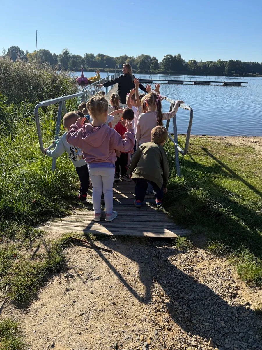 Przedszkolaki z nauczycielką wchodzą na drewniany pomost nad wodą w słoneczny dzień